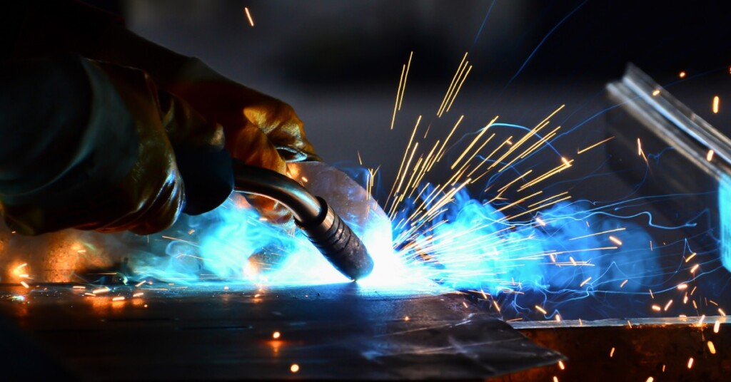 A close up of a welder's hands, torch welding two pieces of metal together, causing sparks and blue flames.
