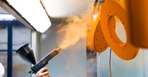 A worker in an all-white coverall suit sprays an orange powder coating on circular metal parts hanging from the ceiling.