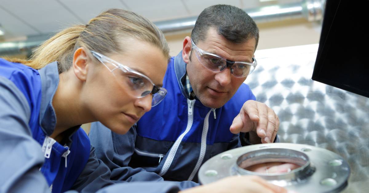 A male and female industrial worker wearing protective eyewear and blue uniforms inspect a polished metal finish.