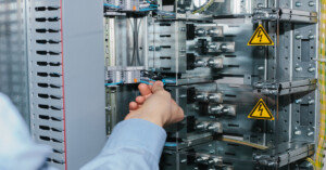 A hand touches blue wires inside an electronic enclosure. Two yellow voltage warning labels are on the cabinet.