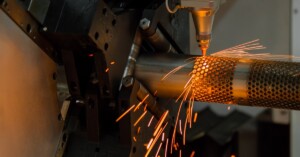 CNC laser cutting machine creates orange sparks while cutting holes in a cylindrical metal workpiece.
