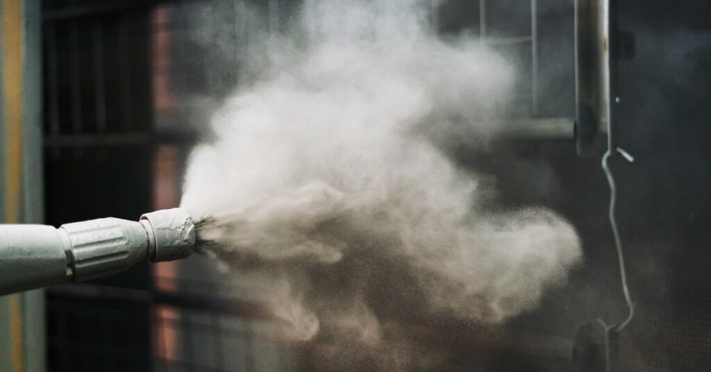 Gray powder coating is sprayed from a nozzle onto a metal lattice part. The nozzle has visible paint debris on its tip.