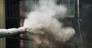 Gray powder coating is sprayed from a nozzle onto a metal lattice part. The nozzle has visible paint debris on its tip.