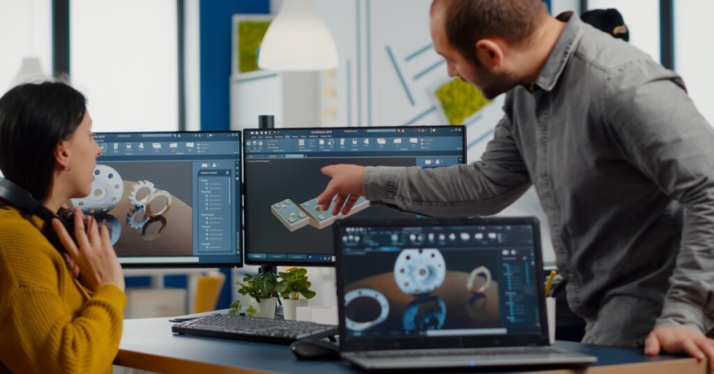 A man points to dual monitors displaying different CAD designs, while a woman sits at a desk looking at them.