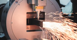 Orange sparks escape from a square metal pipe as a laser cutting machine slices through the surface.