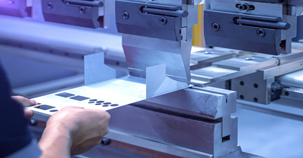 Bare hands hold a flat piece of sheet metal in position, while a CNC machine prepares to bend it into shape.