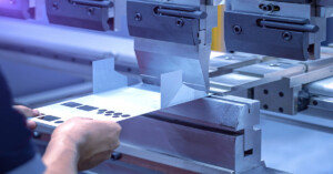 Bare hands hold a flat piece of sheet metal in position, while a CNC machine prepares to bend it into shape.