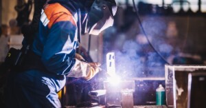 A worker in a black oxygen mask welds a piece of steel, producing visible sparks around the welding point.