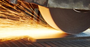 A horizontal grinder machine applies a finish on a metal surface. Sparks fly at the point of contact.