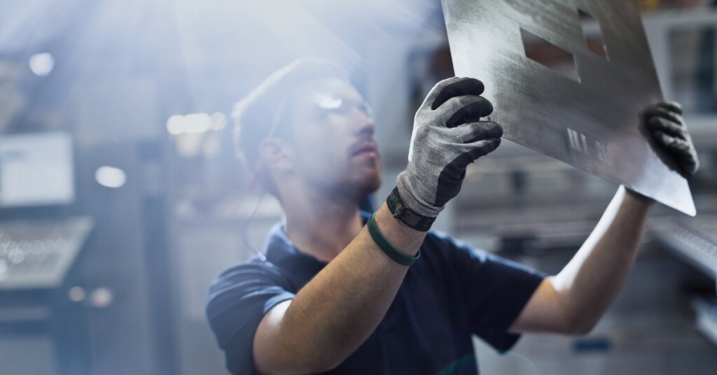 A worker wearing safety gear holds a metal sheet in a bright industrial workshop with blurred machinery in the background.