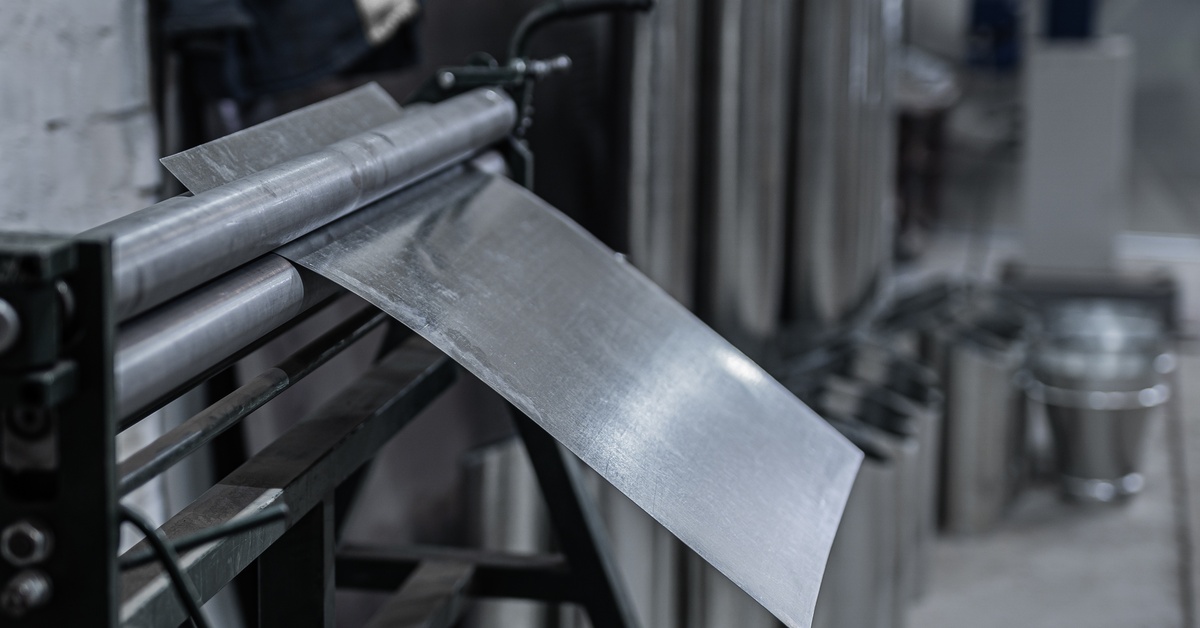 A metal sheet passes through industrial rollers in a factory. There's blurred machinery in the background.