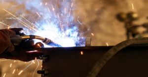 A welder's gloved hand holds a torch, emitting blue light and sparks, to join two metal pieces on a dark surface.