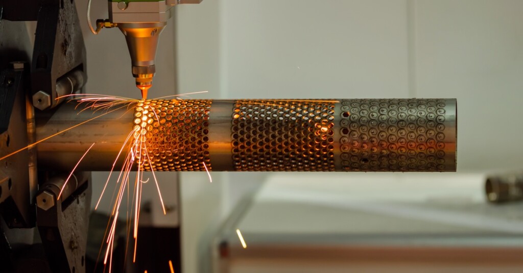 A laser cutting machine emits sparks while perforating a cylindrical metal tube with a precise hexagonal hole pattern.