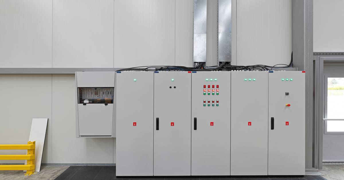 A clean industrial room contains large electrical control panels, indicator lights, cables, and warning labels.