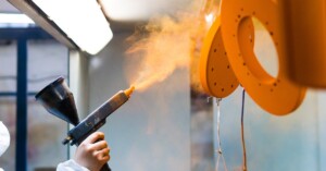 A worker sprays vibrant orange powder onto metal parts in a factory, showcasing an industrial powder coating process.