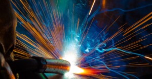 A welder uses a torch, creating bright sparks and blue smoke against a dark background in an industrial setting.
