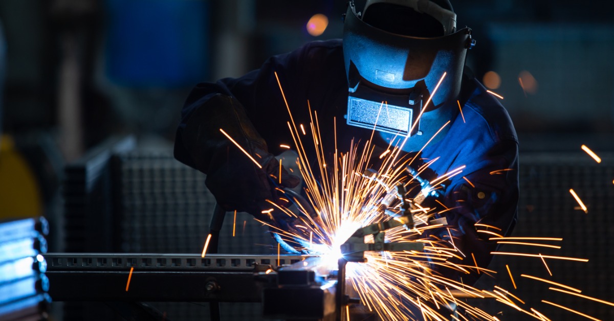 A welder wearing protective gear works on metal, creating bright sparks and a glowing arc in a dark industrial setting.