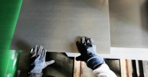 A worker wearing gloves lifts a large sheet metal panel from a stacked pile inside an industrial fabrication facility.