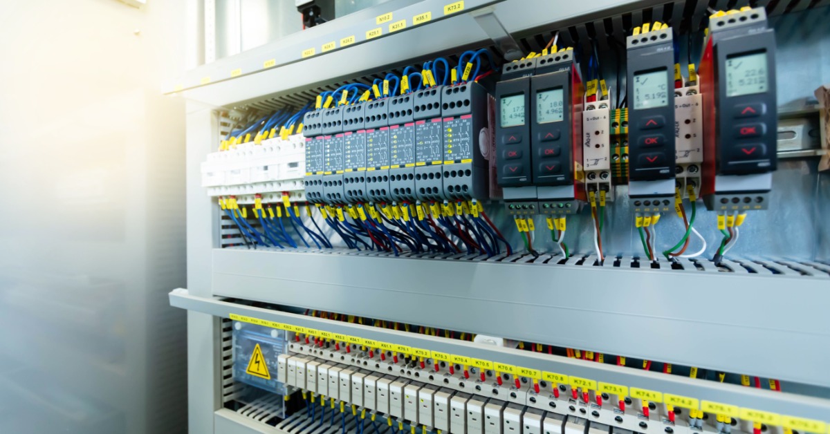 A gray electrical cabinet contains organized circuit breakers, relay switches, and digital displays with color-coded wires.
