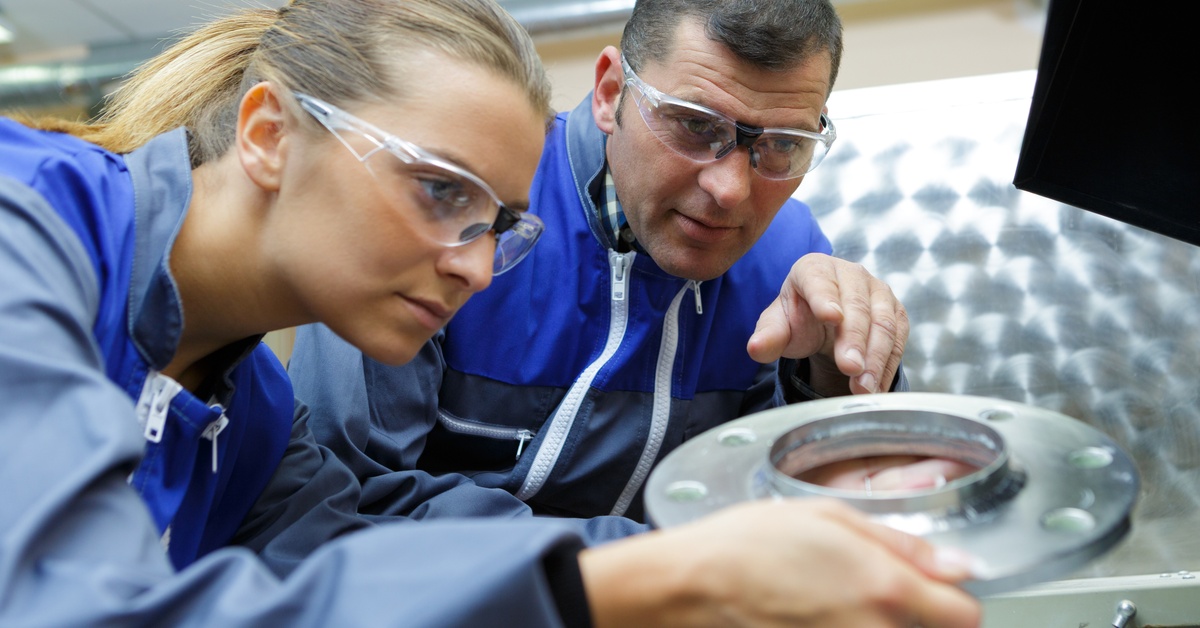 Two engineers wearing safety glasses inspect a metal component near a machining station in a metal fabrication facility.