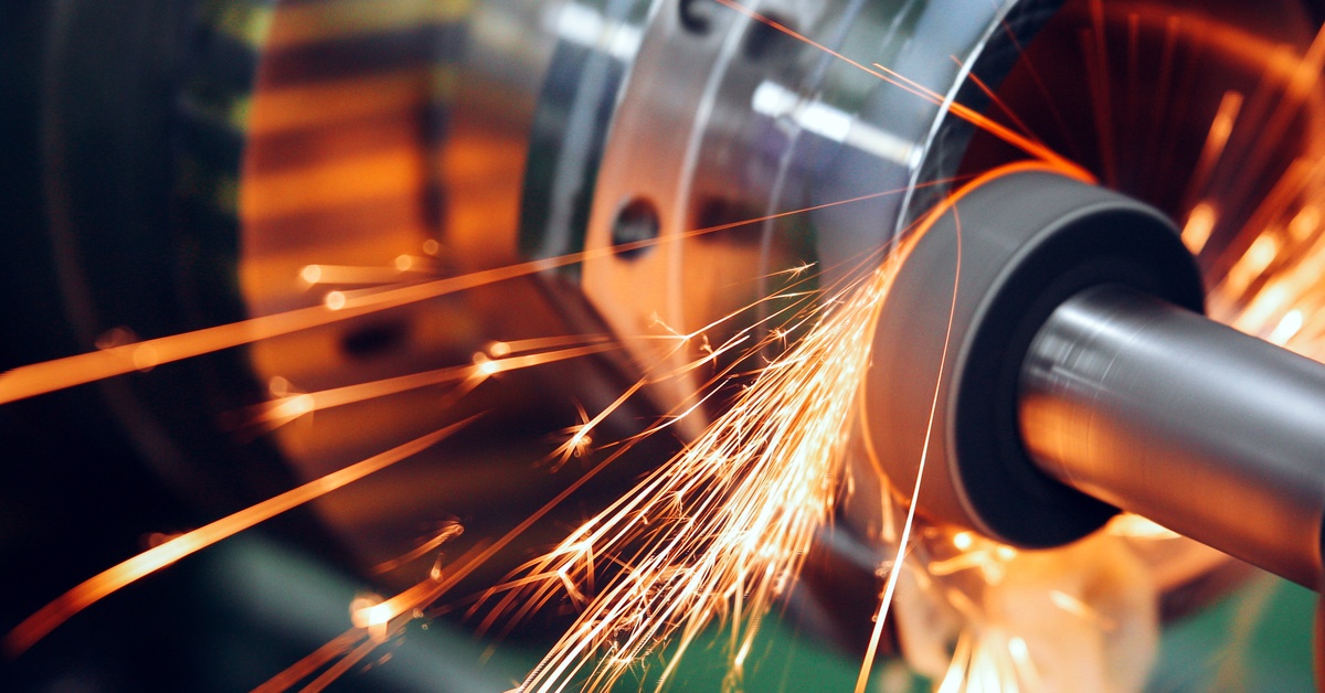 A metal lathe grinds a rotating steel shaft, sending bright orange sparks flying across the workshop floor.
