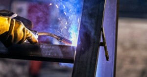 A welding torch creates bright blue sparks and arcs as it fuses a steel beam inside a manufacturing facility.