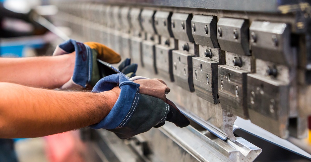A sheet metal fabricator wearing gloves operates a press brake, bending a metal sheet to adhere to the project's specs.