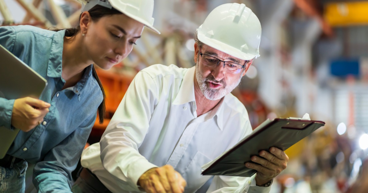 An engineer and a technician holding a clipboard review a metal component, pointing at specific details.