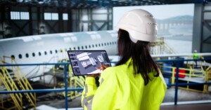 An aerospace engineer looks at a tablet containing aerospace part diagrams while standing beside a hangar.