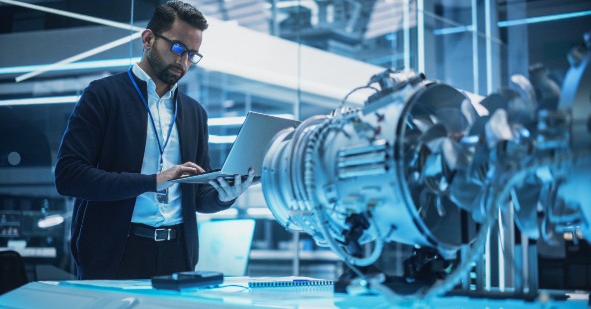 An aerospace technician holding a laptop inspects a turbine engine inside a brightly lit testing facility