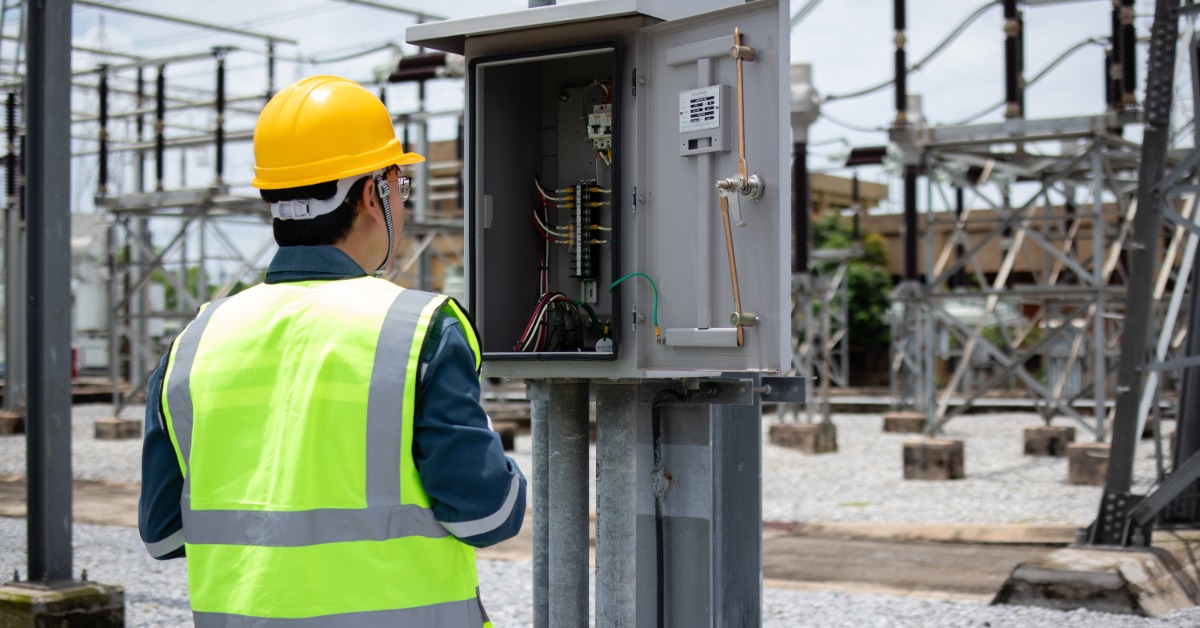 A technician checks an outdoor electrical enclosure mounted on a post in a utility yard with power equipment behind it.