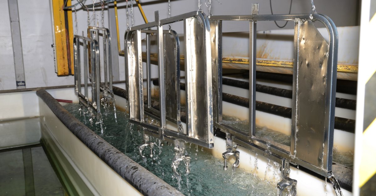 An industrial plating line lifts several metal frames from a bubbling chemical bath inside a factory.