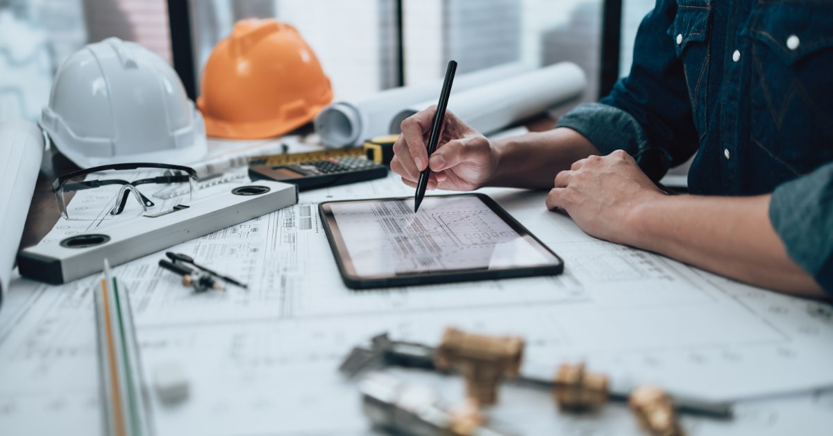 An engineer reviews a technical drawing on a tablet beside blueprints, safety glasses, and construction tools.